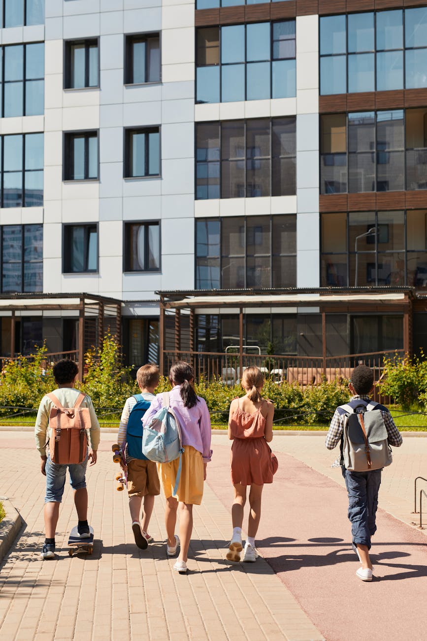 Five children with school backpacks walk toward a school building on a sunny day, seen from behind.