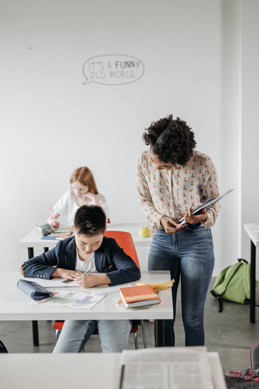 A teacher leans over to help a student working at his desk in a bright classroom, another student visible in the background.